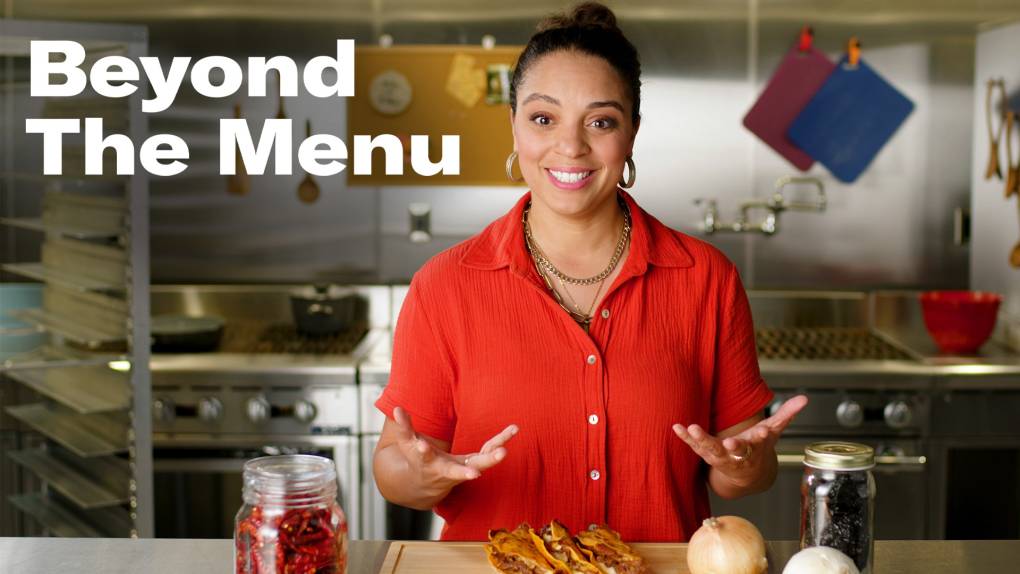 A woman in a red shirt is standing in a commercial kitchen and is excited to try the tacos that are right in front of her.