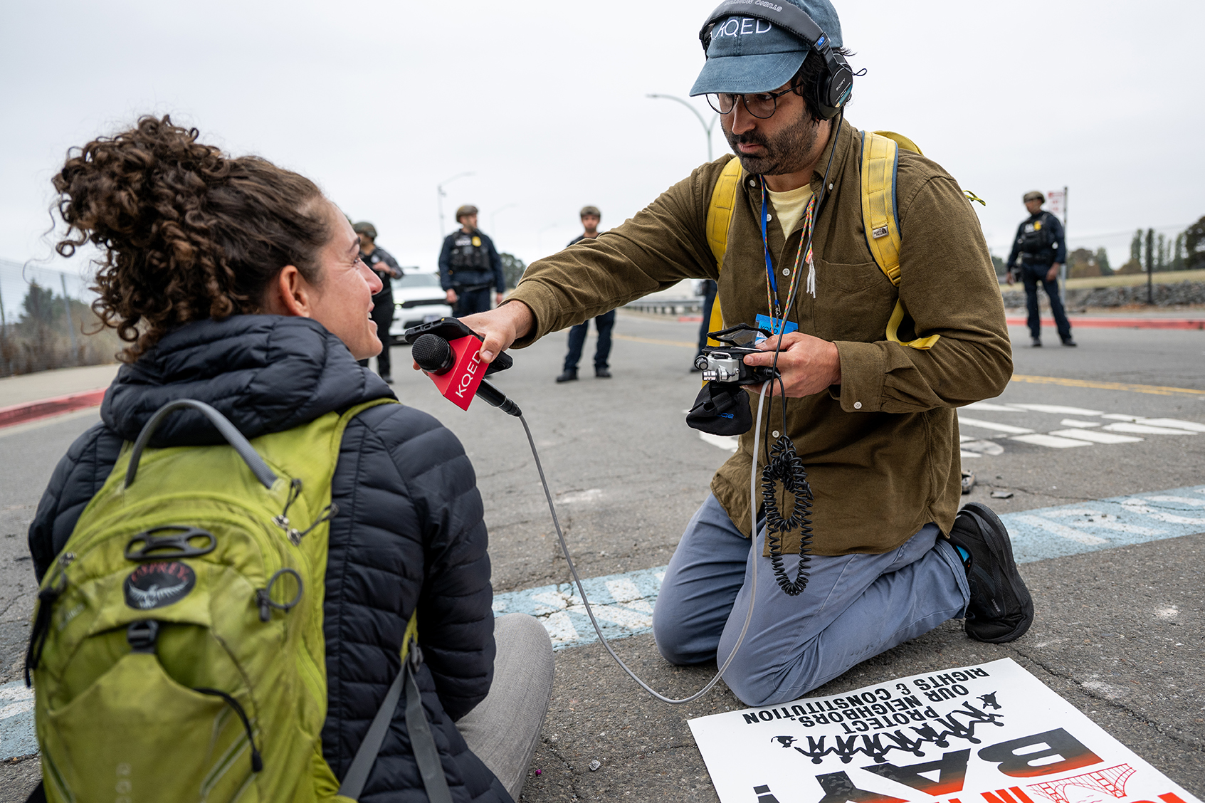 Reporter Azul Dahlstrom-Eckman wears a cap labeled “KQED,” over-ear headphones, glasses, and a yellow backpack kneels on a paved street while conducting an interview with a protester in front of the entrance to a U.S. Coast Guard base in Oakland on Oct. 23, 2025. He holds a red KQED-branded microphone toward the female protester seated on the ground, who wears a dark puffer jacket and a bright green backpack. Dahlstrom-Eckman also holds a small audio recorder with cables attached. In the background, several police officers stand spaced out across the road, slightly out of focus. A protest sign with partially visible text lies on the ground in the foreground near the reporter’s knee. The sky is overcast, and the overall scene suggests an on-location interview during a public demonstration or street closure.
