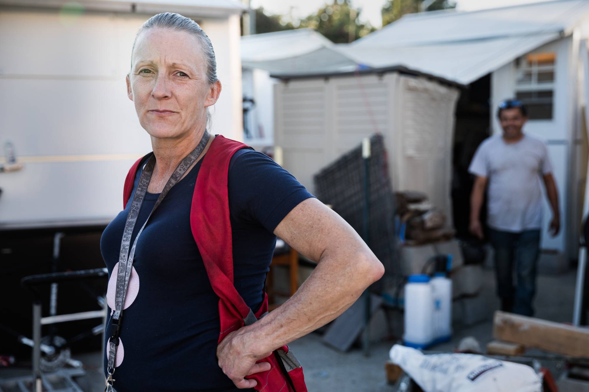 Tammy, a former resident of Oakland's East 12th Street Parcel, stands outdoors in the parcel, facing the camera with a steady, confident expression and one hand resting on her hip. She wears a dark short-sleeve shirt and a red vest over her shoulders, along with a lanyard hanging around her neck. Her hair is pulled back, and natural light highlights her face.
Behind her are small structures or sheds, a canopy or tarp, and various items such as containers, tools, and stacked materials. A second person is visible in the background, slightly out of focus, walking through the space. The setting suggests a lived-in, transitional outdoor environment, and her posture conveys resilience and self-possession.