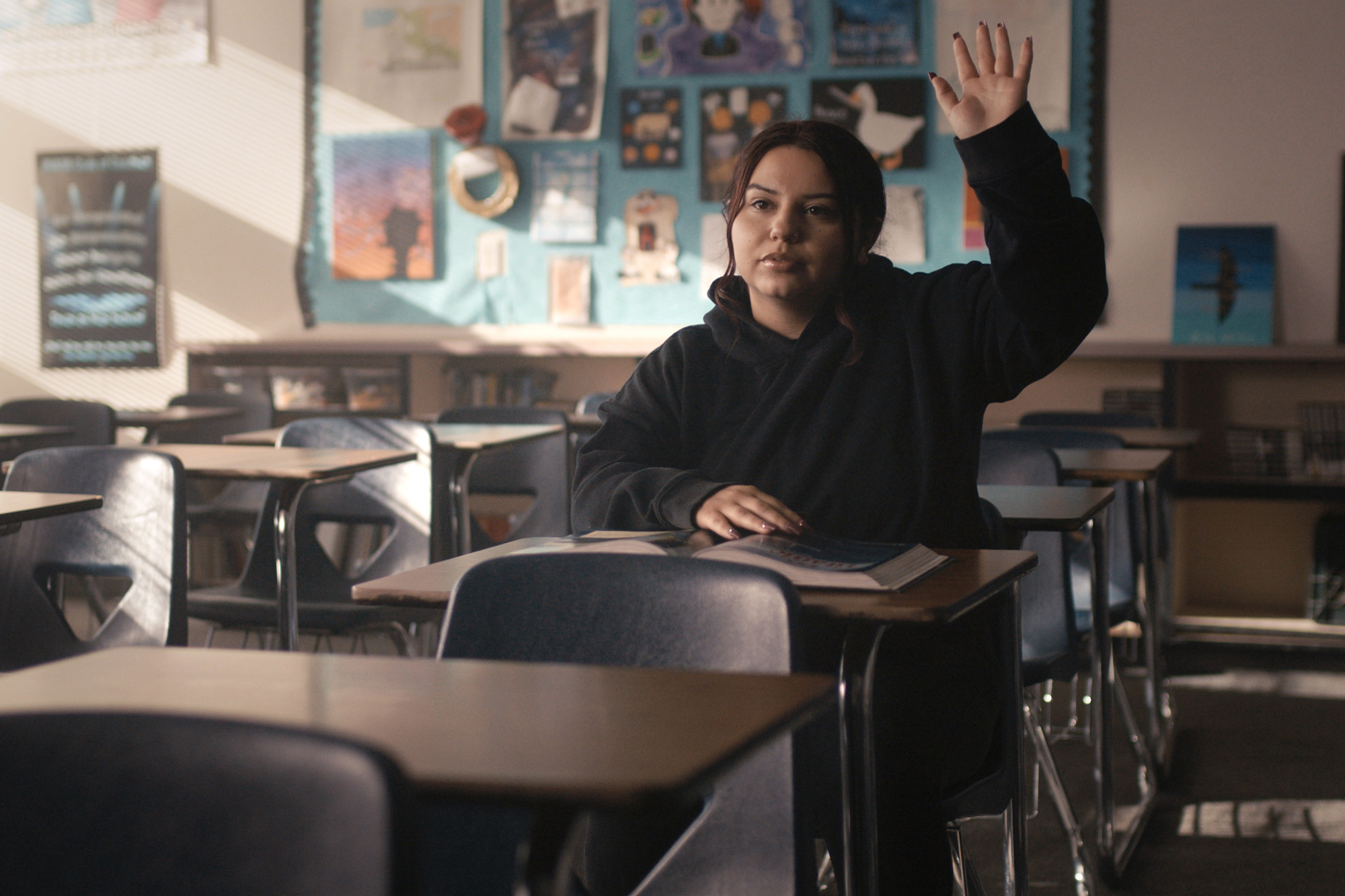 Emily Huizar, one of seven Deer Valley High School students featured in The Class, sits alone at a desk in a quiet classroom. She wears a black hoodie and raises her right hand, appearing ready to answer a question. A notebook lies open on her desk. The classroom around her is mostly empty, with rows of desks and chairs extending into the background. Sunlight filters through window blinds on the left, creating striped patterns of light and shadow across the room. Behind her, a bulletin board displays colorful posters and artwork, adding contrast to the otherwise muted tones of the classroom.