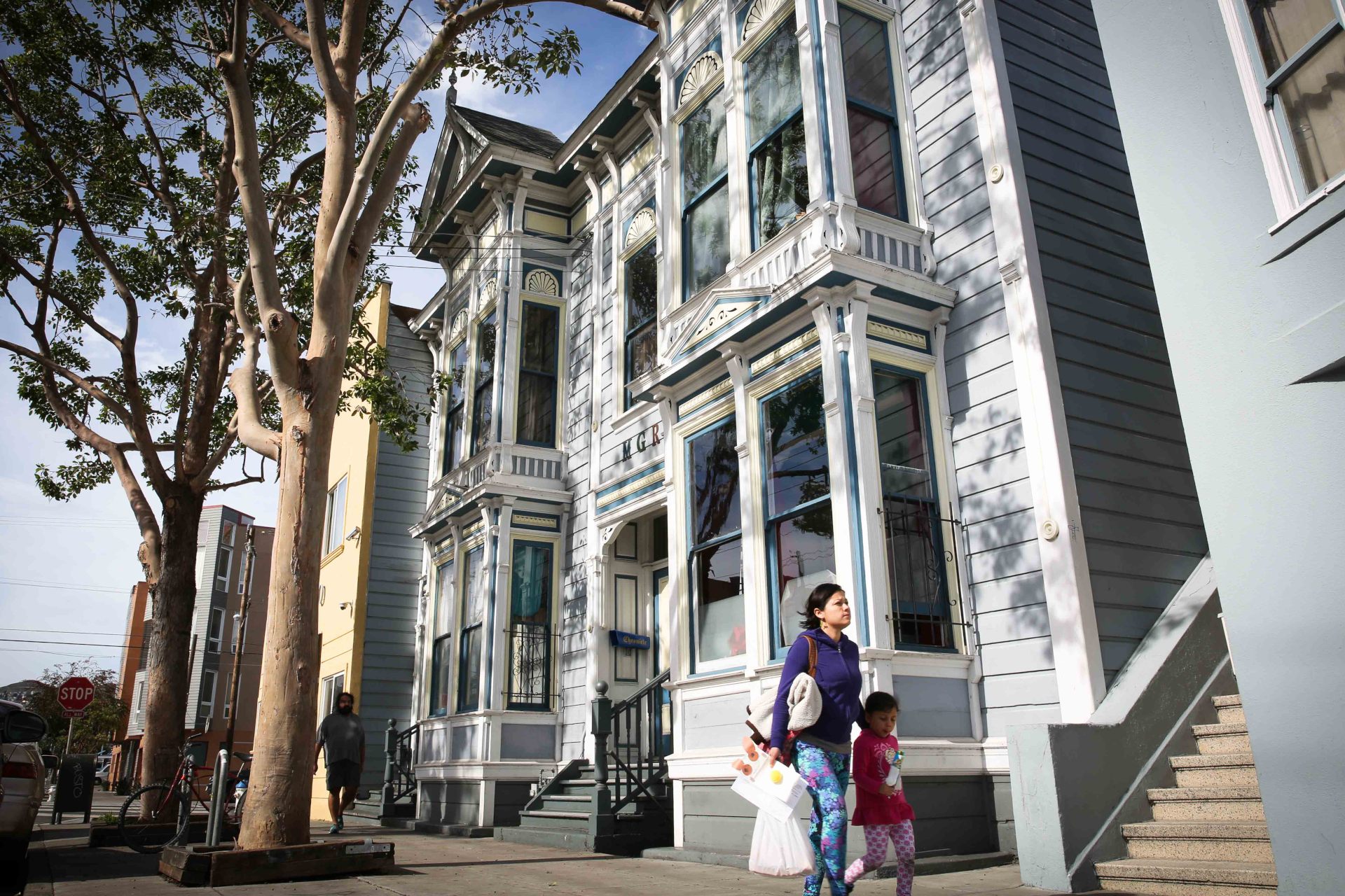 A woman holding the hands of two small children walks down the street past a large home.
