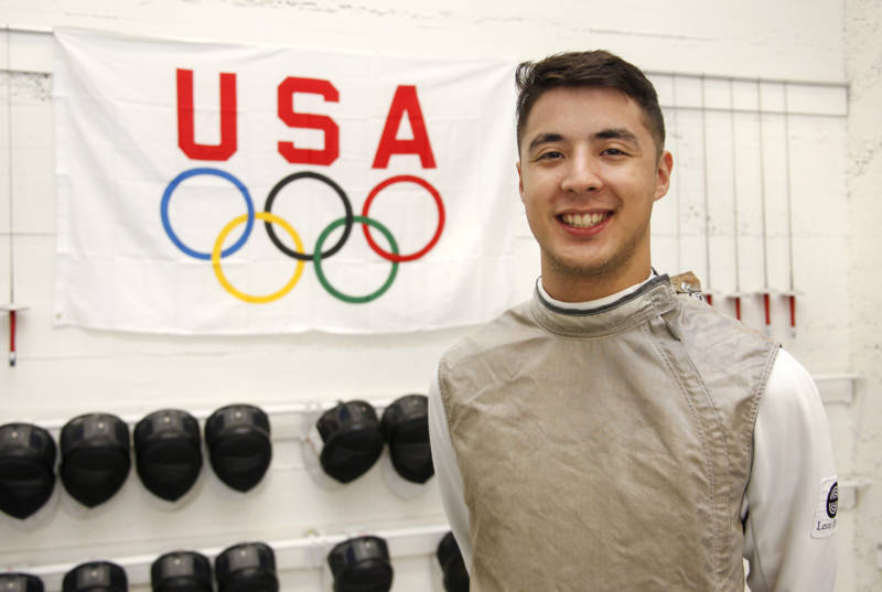A man wearing fencing gear stands in front of an Olympic USA flag with fencing helmets underneath.