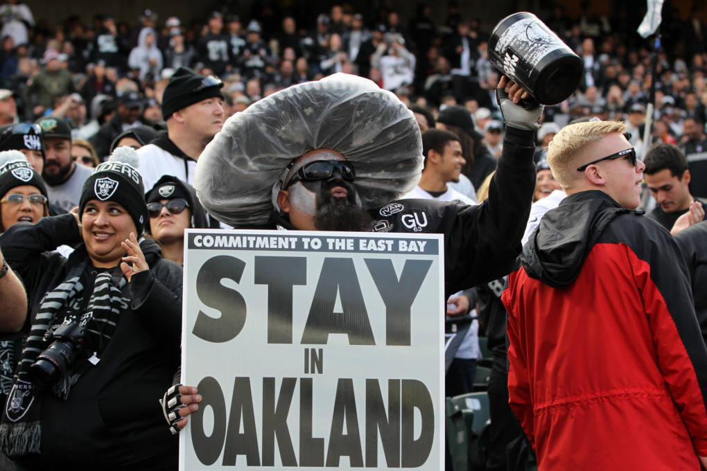 A person wearing black and white make-up and clothing holds a sign that reads "Stay in Oakland."