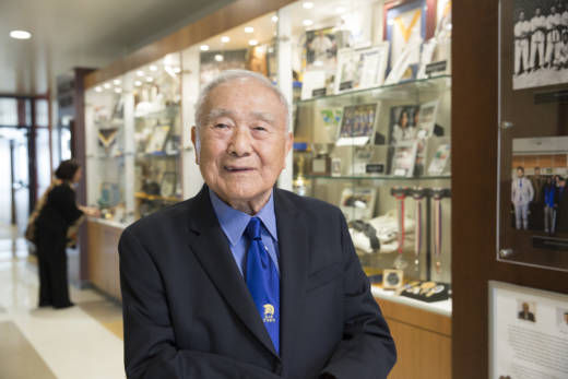 Coach Yoshihiro "Yosh" Uchida standing in front a case of judo medals outside the dojo in a building named after him at San Jose State.