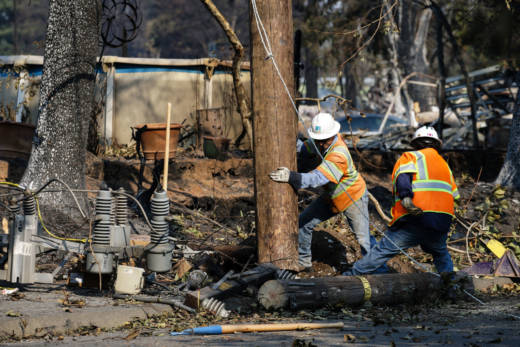 Two energy workers dressed in safety gear and neon colors hold a tree amid a fire-damaged neighborhood.