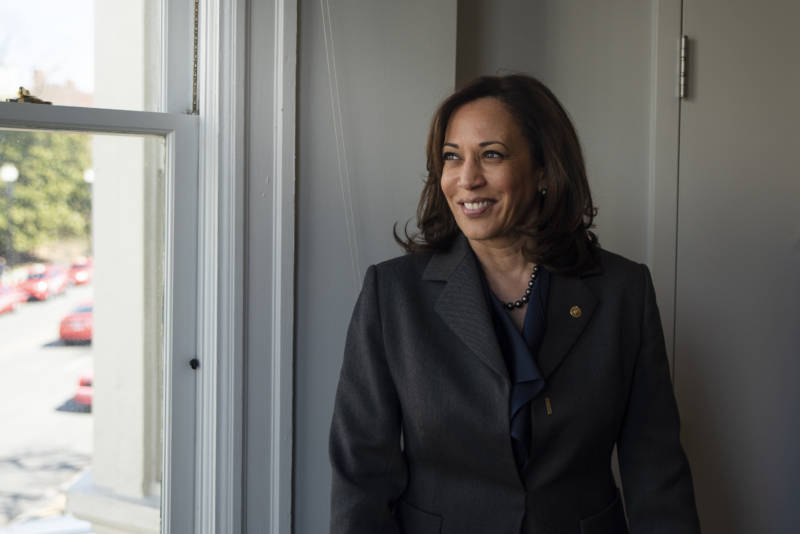 A Black woman wears a dark-colored suit while smiling and staring out a window to her right.