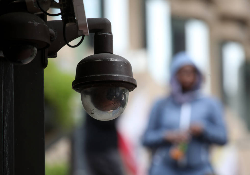 A video surveillance camera is seen hanging with a blurred person in the distance.
