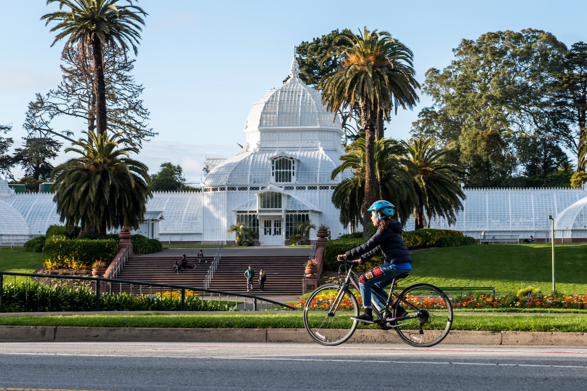 A person on a bike rides past a big white building surrounded by green grass and tall trees.