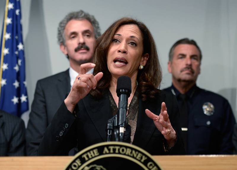 A woman wearing a business suit gestures with her hands while standing at a podium. Two men are behind her.