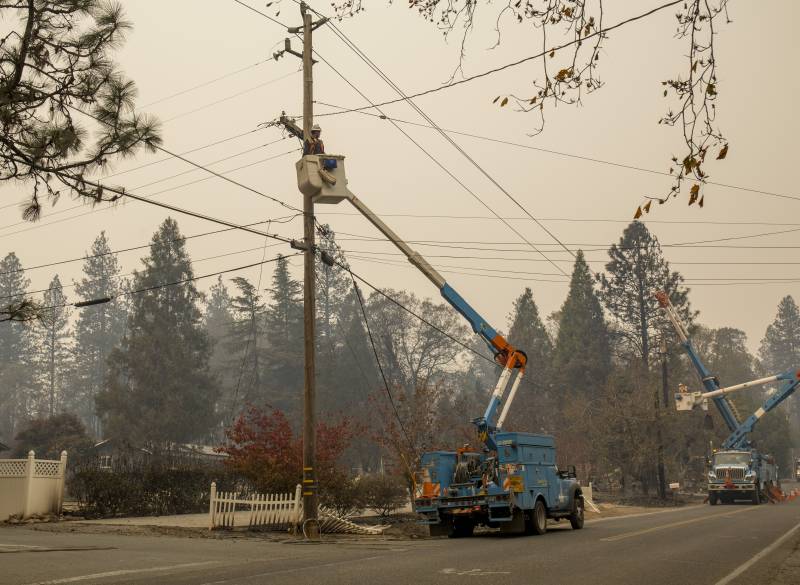 Two cranes hoist up two workers toward electrical lines. In the background is a smoky sky and trees.