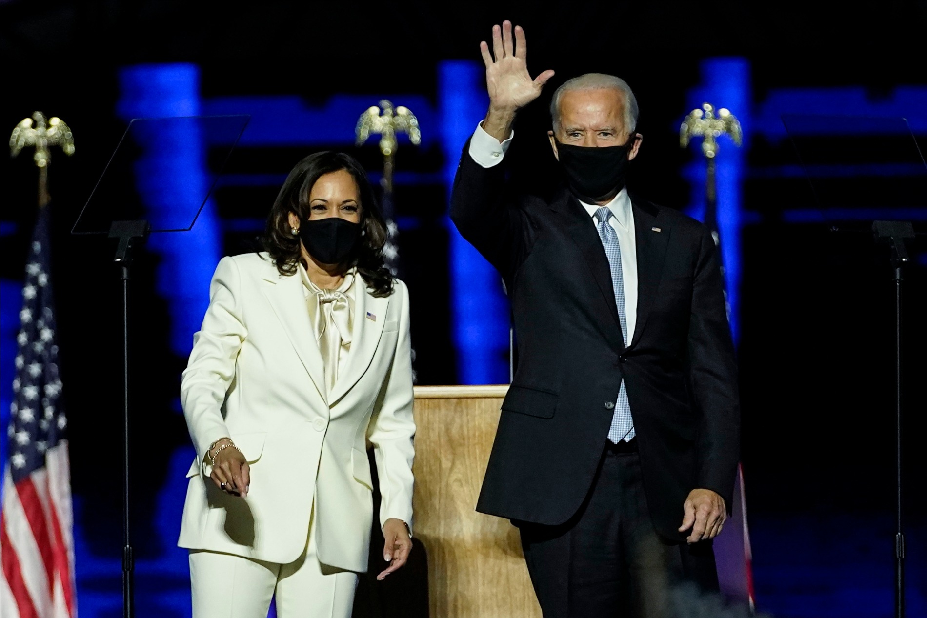 A woman wearing a white suit and face mask stands next to a man in a black suit and face mask that's waving.