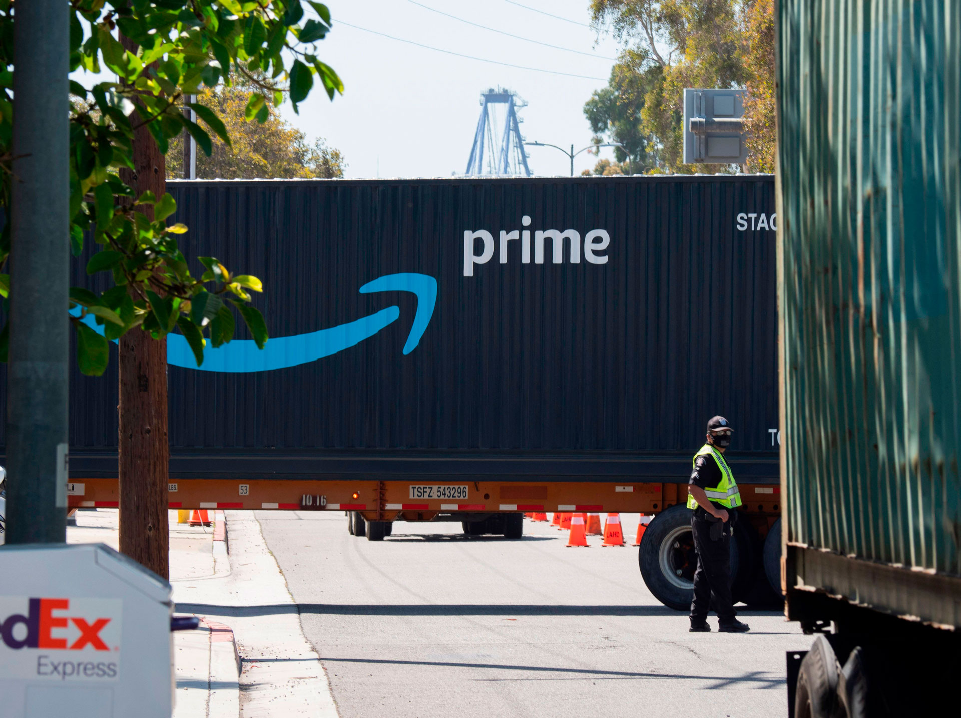 A shipping container with an arrow pointing to the word "prime" is seen on the street. One person in a safety vest stands to the right side of the container.