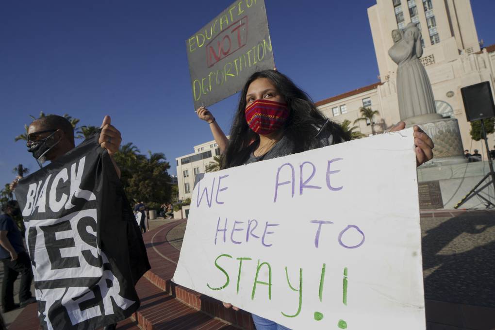 Protesters hold signs saying, "We are here to stay!" "Black Lives Matter" and "Education, Not Deportation."