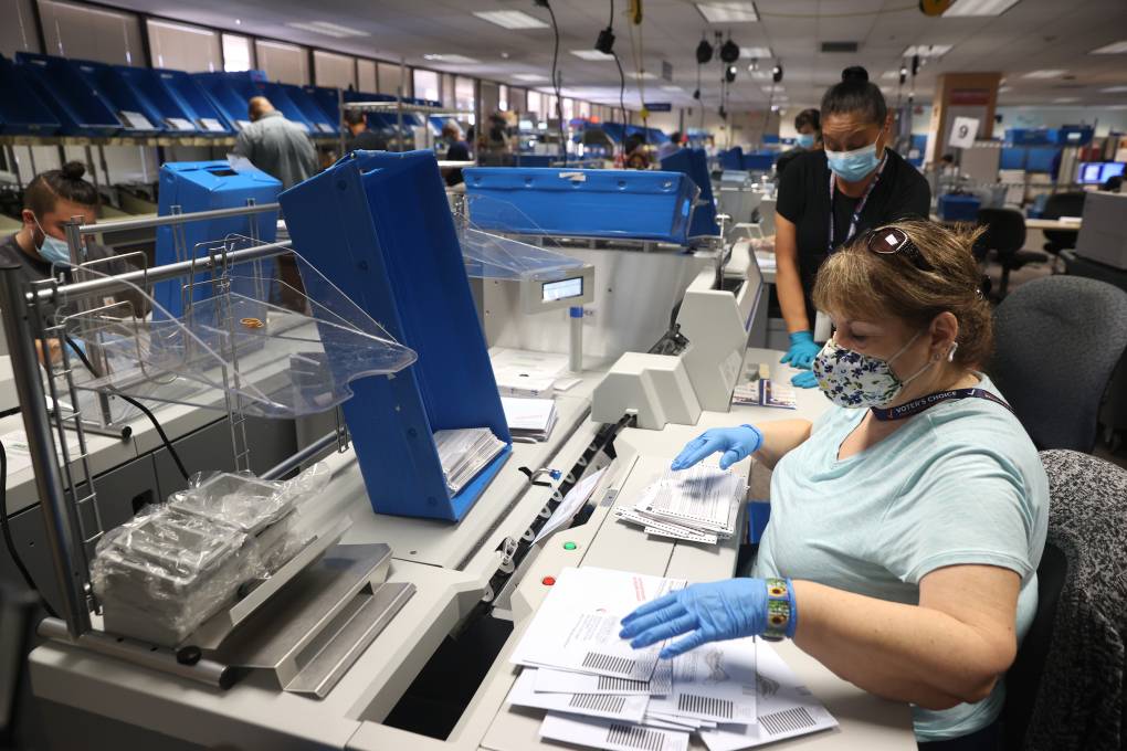 A woman wearing a face mask and gloves sits at her desk with dozens of envelopes in front of her. Other staff members work nearby.