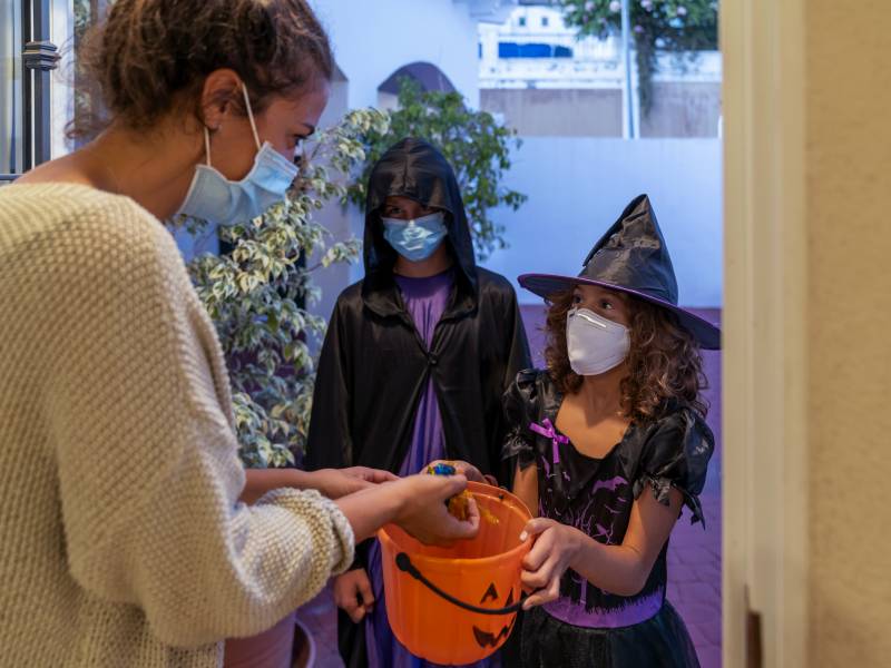 Children dressed in Halloween costumes and face masks accept candy from an adult at her front door.