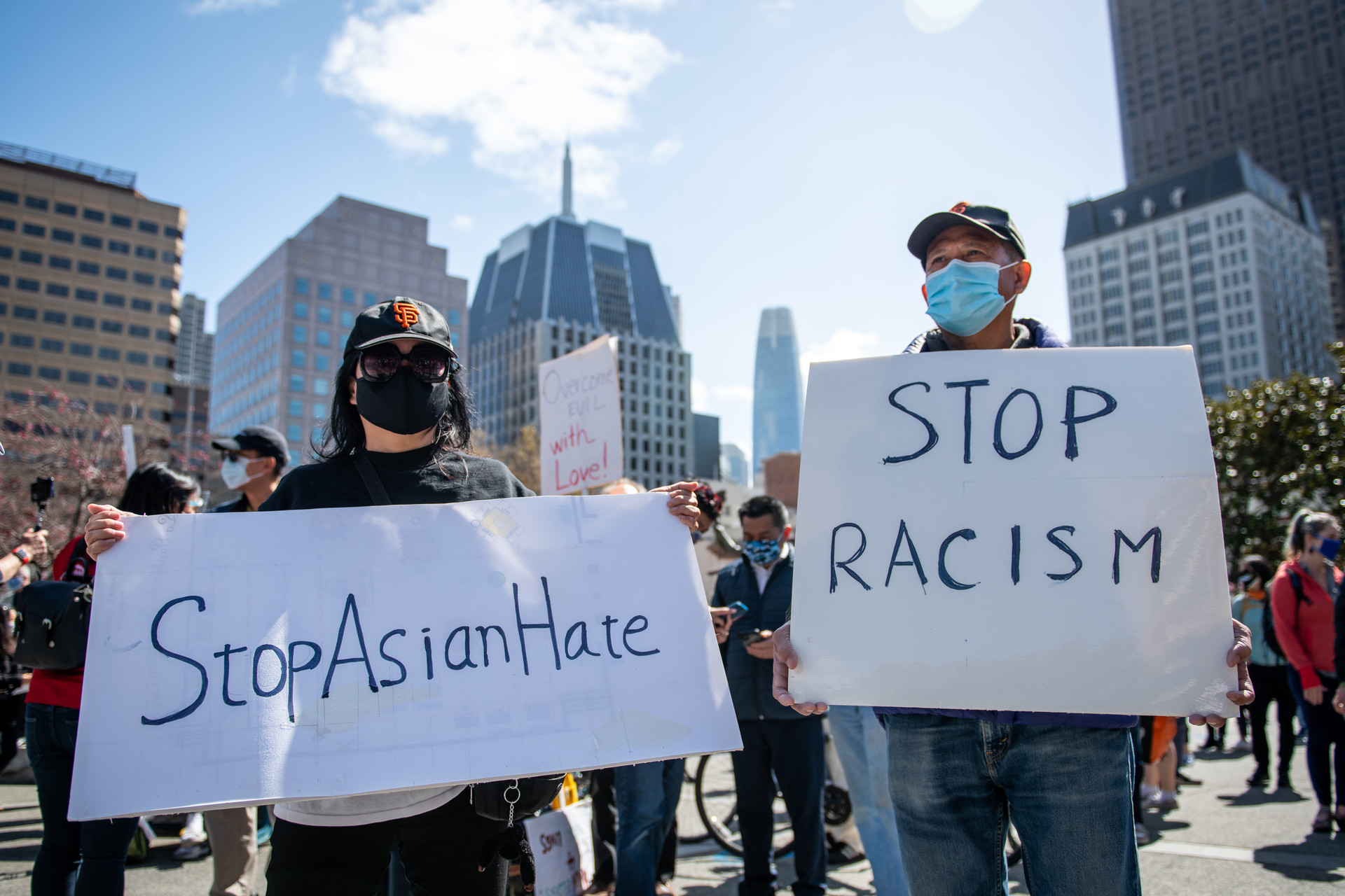 Two people wearing black masks in a crowd hold signs that say "stop Asian hate" and "stop racism."