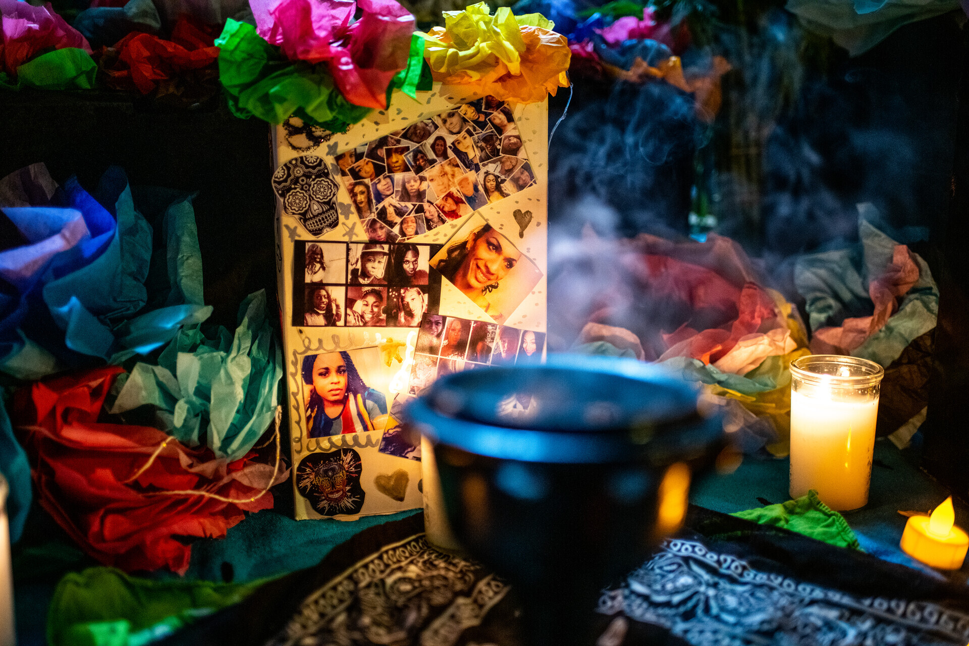 A collage of photos with pink, green, and yellow paper flowers stands next a small mortar that carries burning sage. A few candles are nearby.