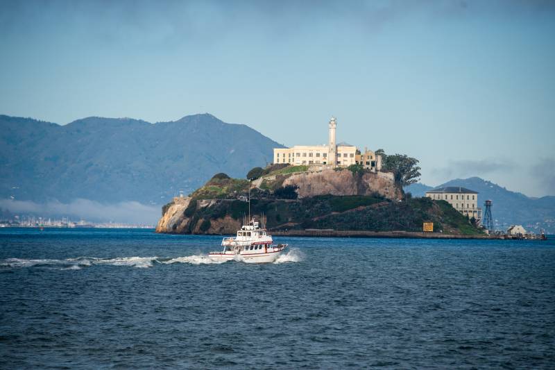 An island with a prison and a lighthouse visible atop it on a sunny day. A ferry passes by in the foreground.
