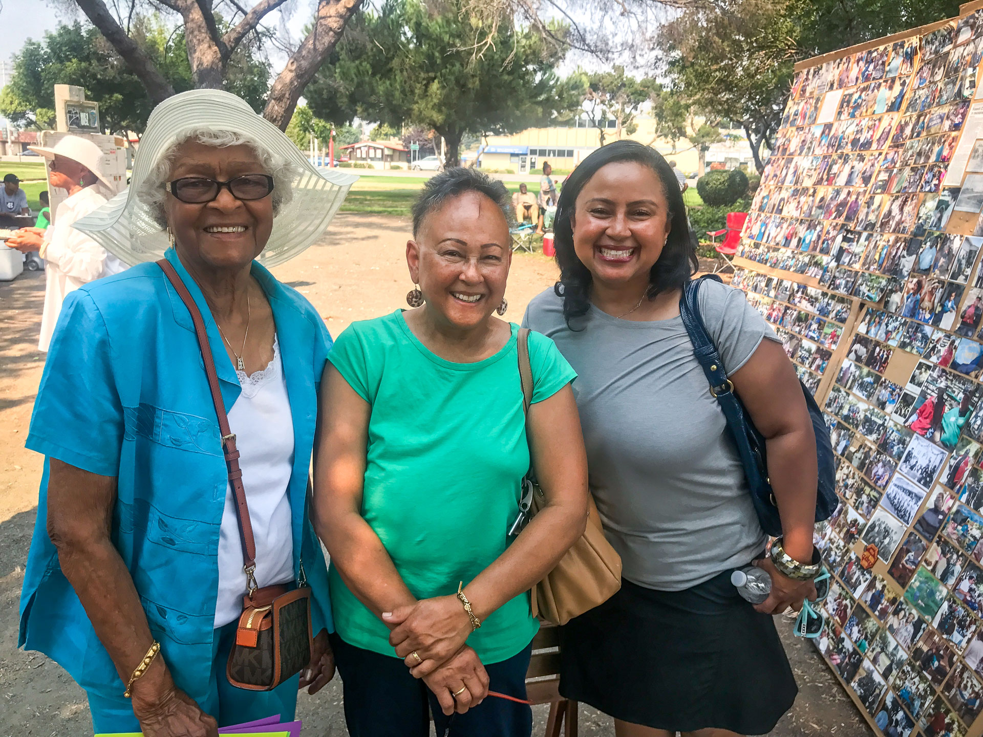 Three women stand together. The woman on the left is wearing glasses, a turquoise sweater with a brown purse. The woman in the middle with her hand holding her wrist is wearing a green shirt and light brown bag. The woman on the right is wearing a grey shirt with a black purse on her left shoulder.