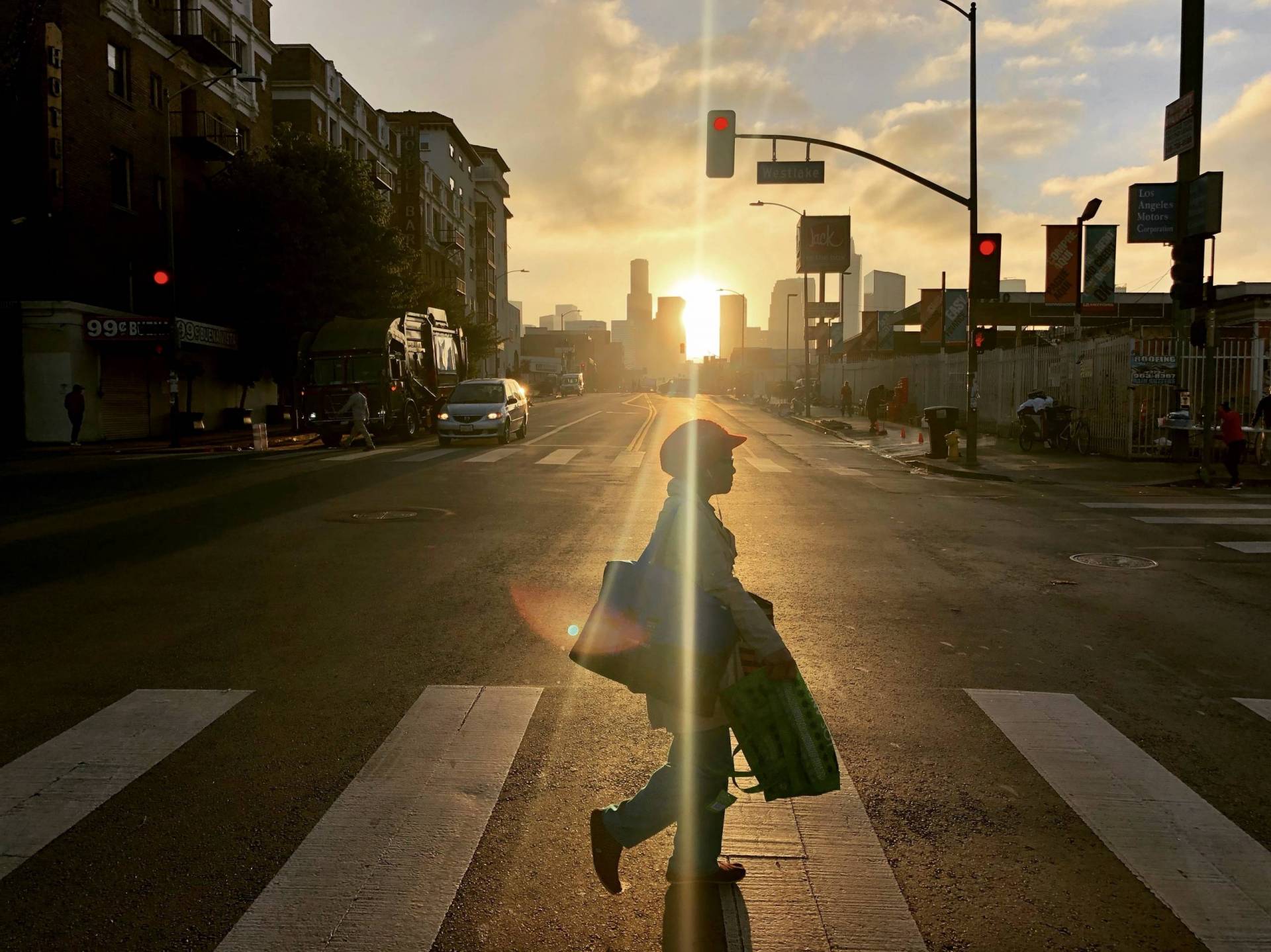 A lone person crossing a broad street, with the sun rising behind her.