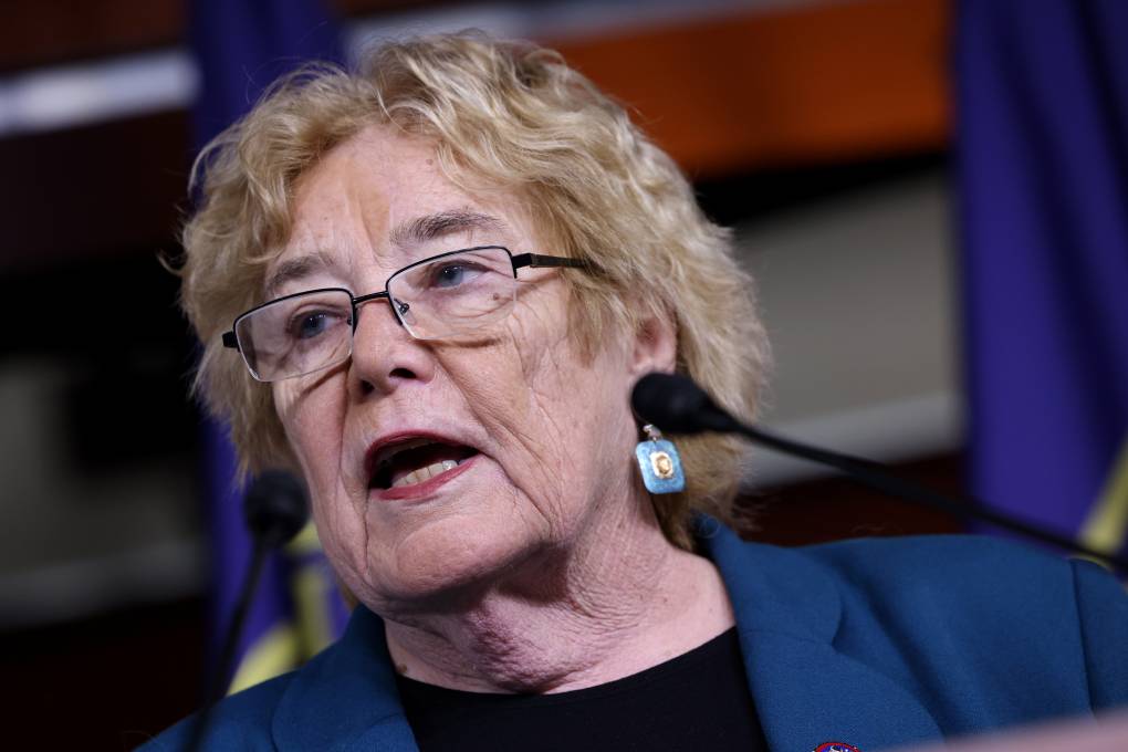 A close-up photo of an older white woman with blonde hair wearing glasses and a blue suit jacket. Two blurry microphones can be seen in front of her.