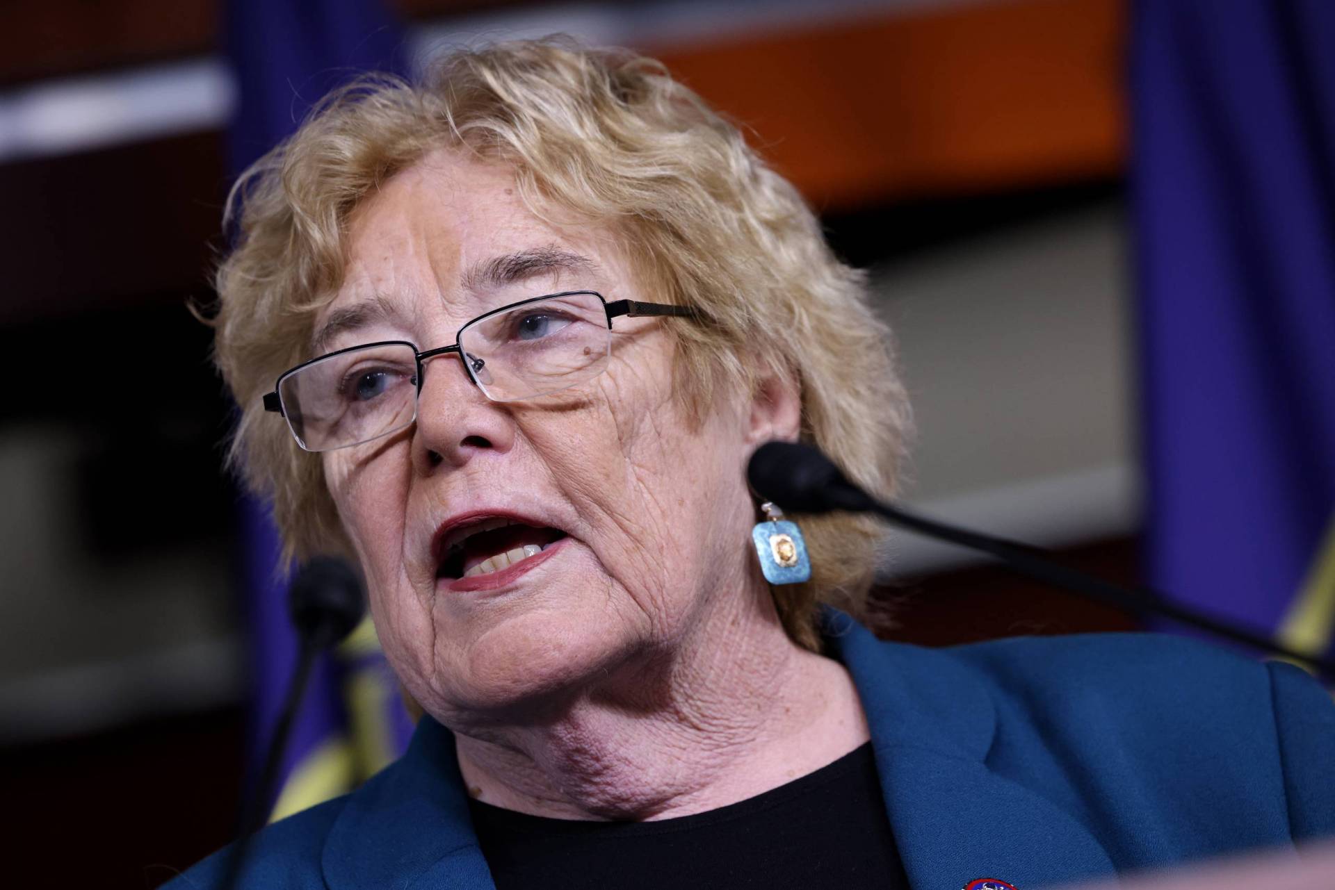 A close-up photo of an older white woman with blonde hair wearing glasses and a blue suit jacket. Two blurry microphones can be seen in front of her.