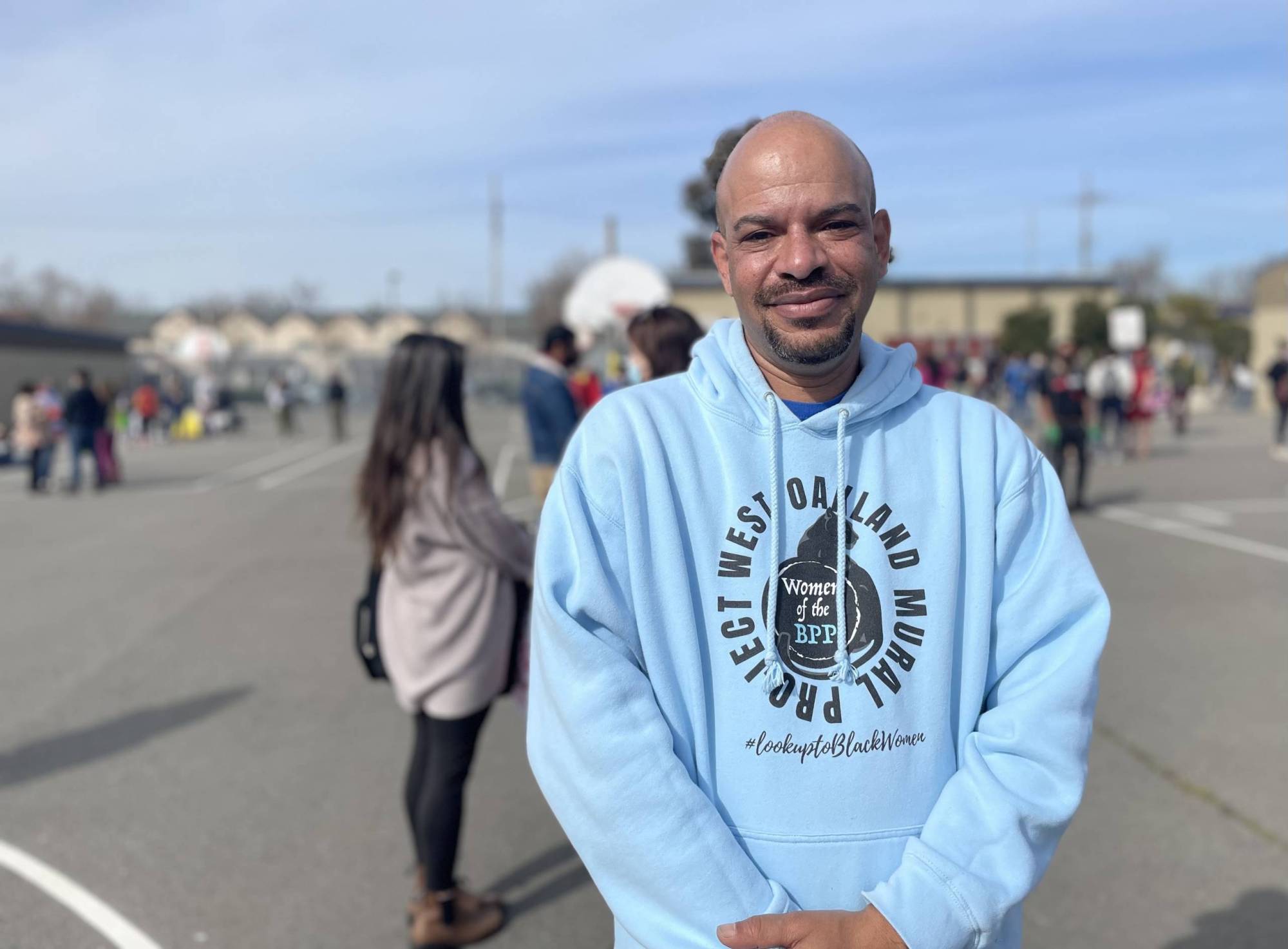 A bald man in a light blue hoodie stands for a portrait. Behind him a scattered crowd stands on blacktop at Prescott School.