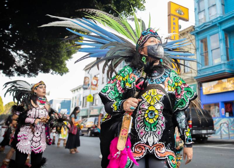 A person wearing a beaded and multicolored blouse stands in the foreground, wearing a mask. A plume of feathers in black, blue, yellow and white fan out behind their head.