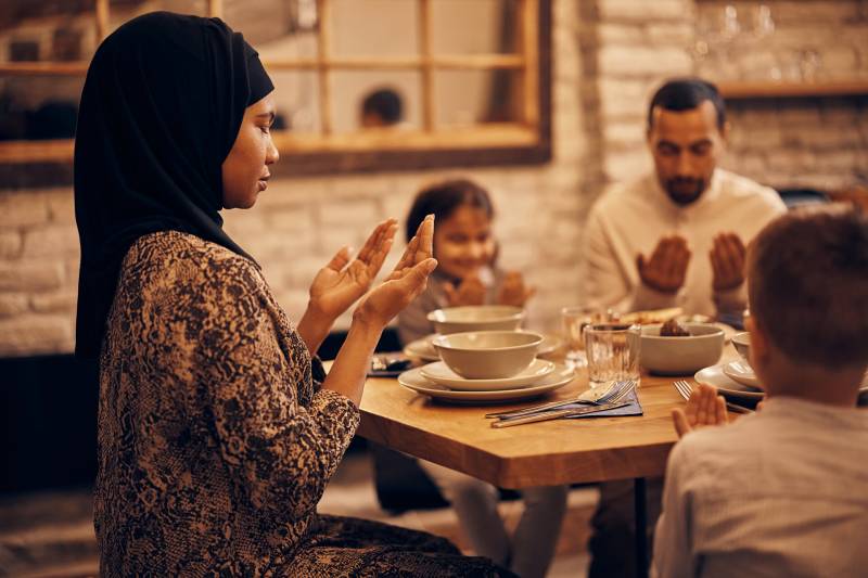 A Muslim family sits around a dining table, eyes closed, palms lifted, face-up. On the left, at the head of the table, sits a woman wearing a black hijab and a patterned dress. Two children sit on either side of her with their hands in the same position; we see the back of a small, close-cropped head to her right, and to her left sits a young girl with shoulder-length dark hair, smiling with her eyes closed. Beside the girl sits a man wearing a white button-up shirt. The table is set neatly with white bowls and plates and clear glasses. The wall behind the table is white-painted brick, inset with a wall-size mirror. The overall tones in the photos are cream and sepia.
