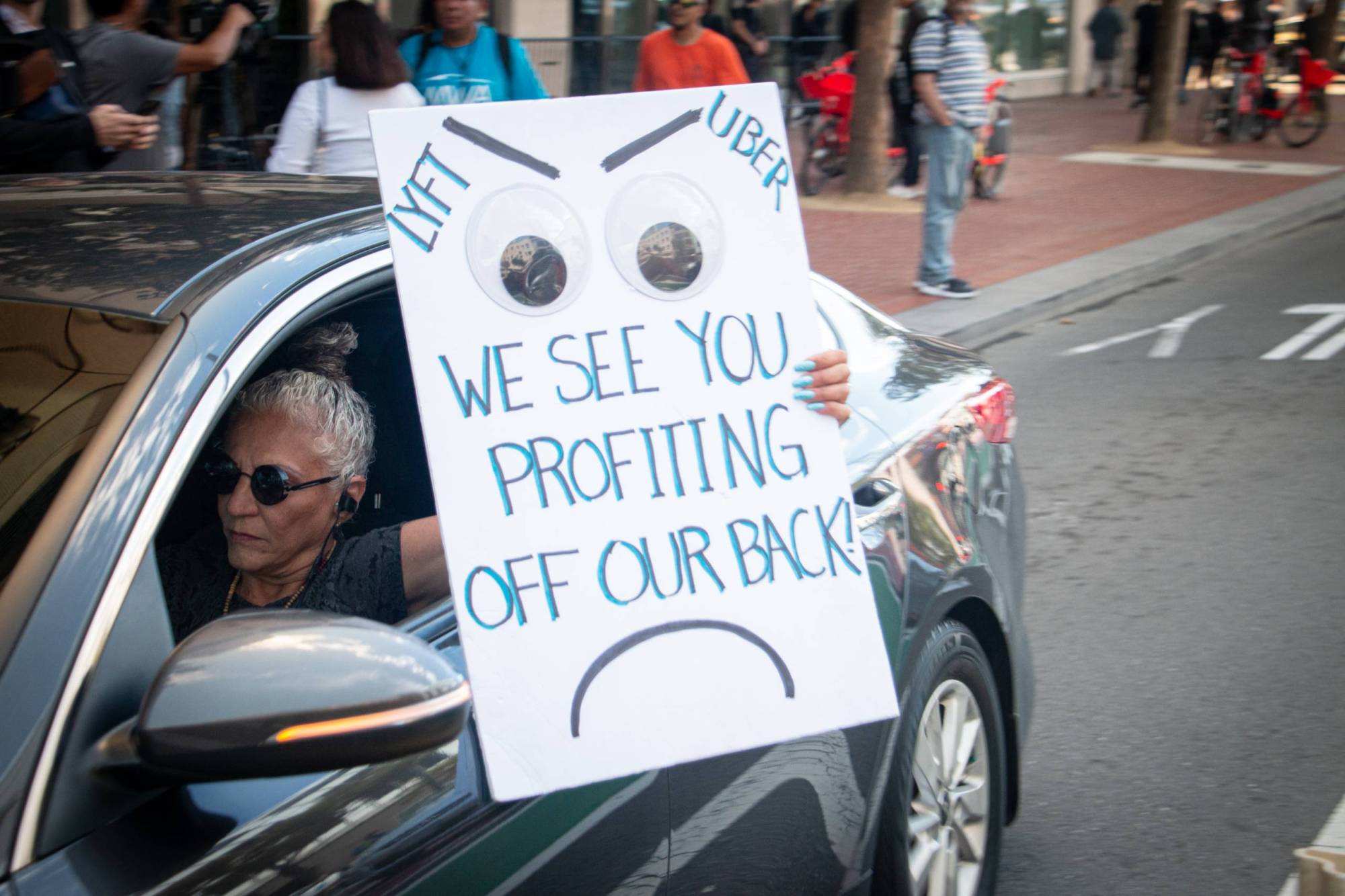 A woman wearing sunglasses drives a car while holding a sign that says: "Lyft and Uber we see you profiting off our back!"