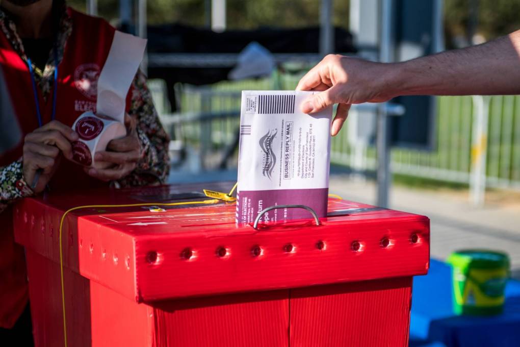 A hand places a ballot into a red ballot box.
