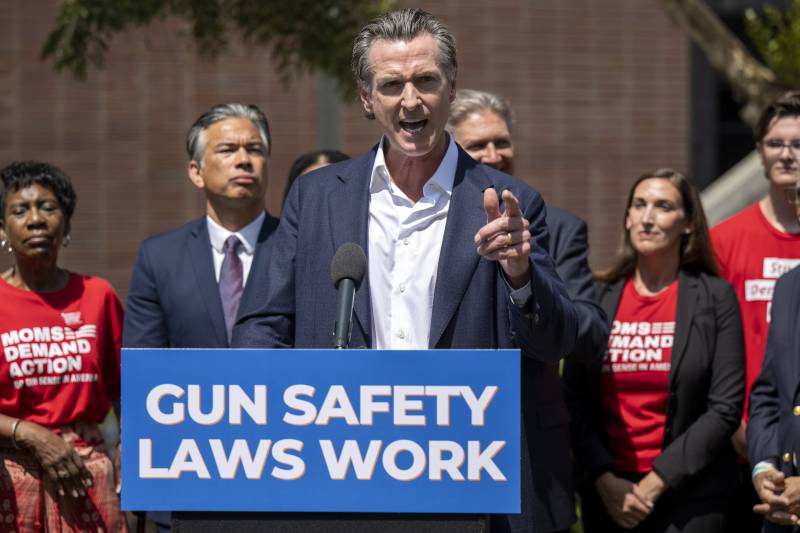 A white man wearing a business suit stands at a podium pointing a finger with people standing behind him.