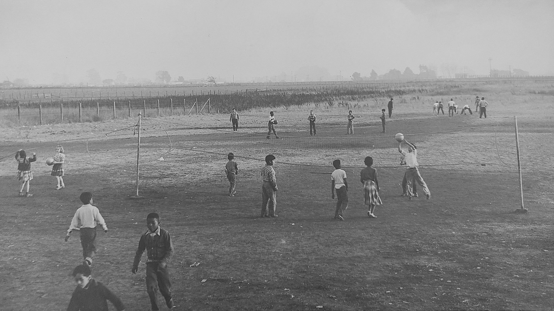 Black and white photograph of kids playing volleyball by an empty field. Some low buildings rise up in the background