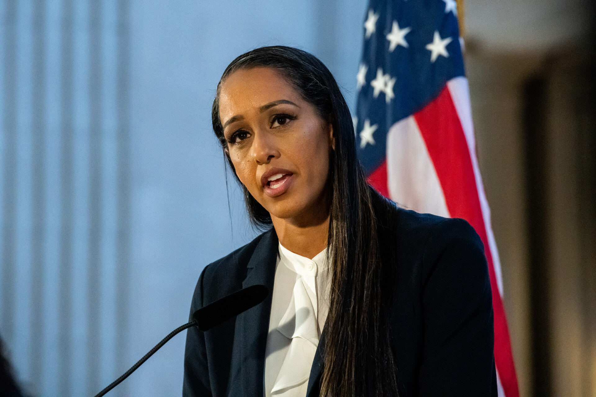 A woman wearing a business suit stands in front of a microphone with the American flag in the background.