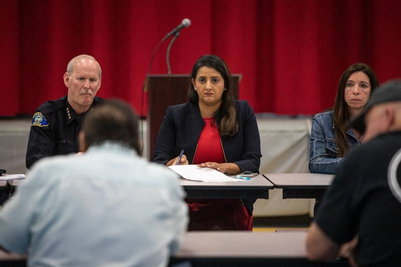 a white man sites to the right of an Indian woman with another woman sitting to the left as they sit behind a table and listen to a man speak.