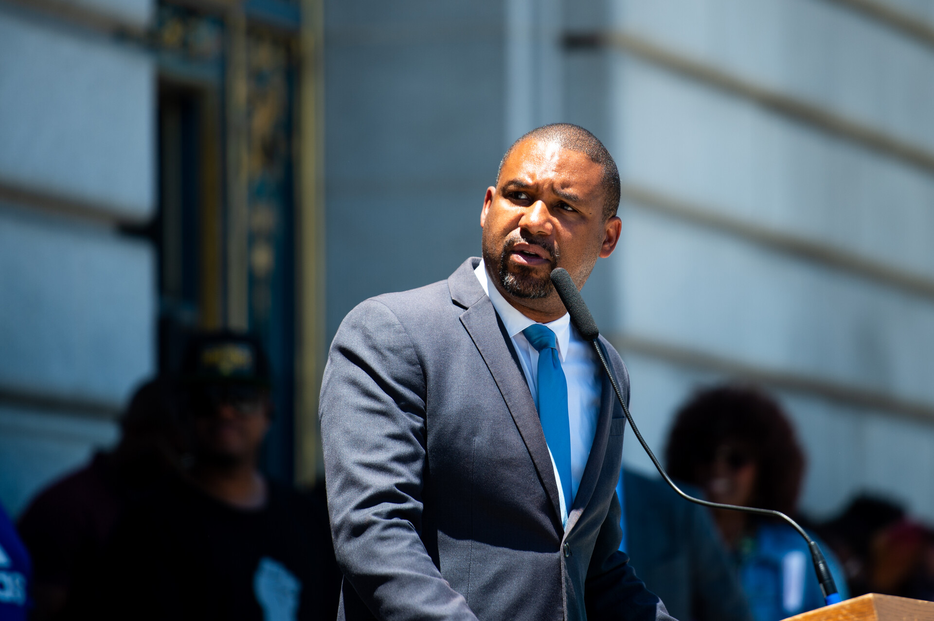 A man in a suit with a blue tie speaks at a rally in front of a microphone.