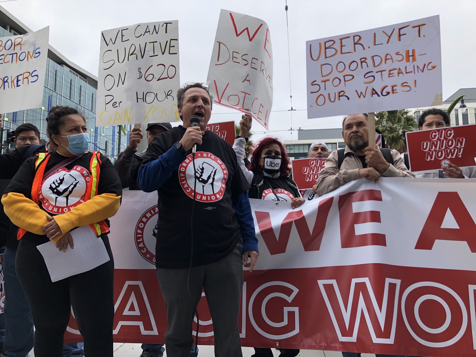 A man with a microphone speaks while protestors holding signs rally behind him.