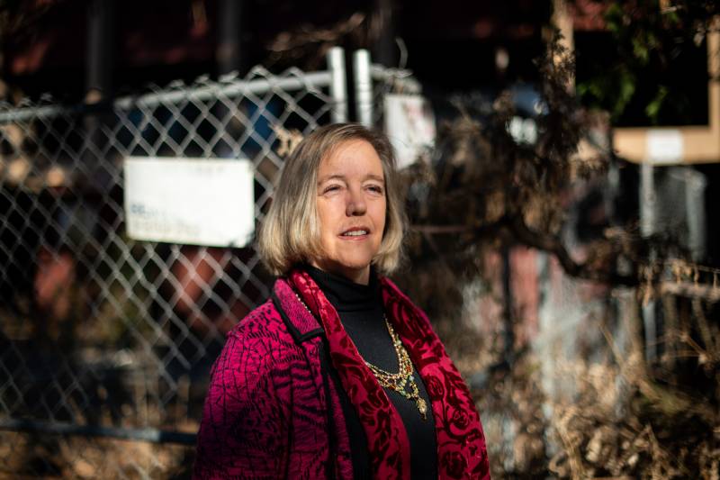 A middle-aged white woman with blond hair wearing a crimson coat and an ornate necklace stands outside in front of a fence.