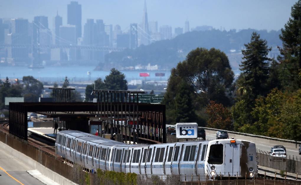 A BART train pulls out of an above-ground station. Cars whiz by alongside and the city of San Francisco looms in the background.