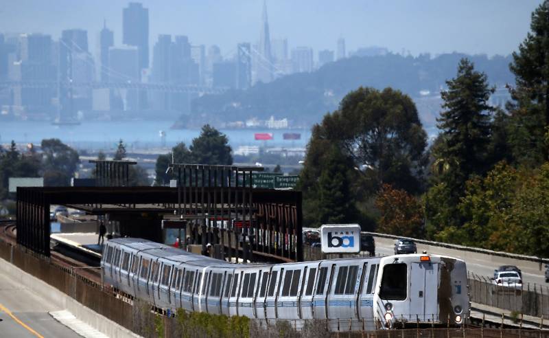 A BART train pulls out of an above-ground station. Cars whiz by alongside and the city of San Francisco looms in the background.