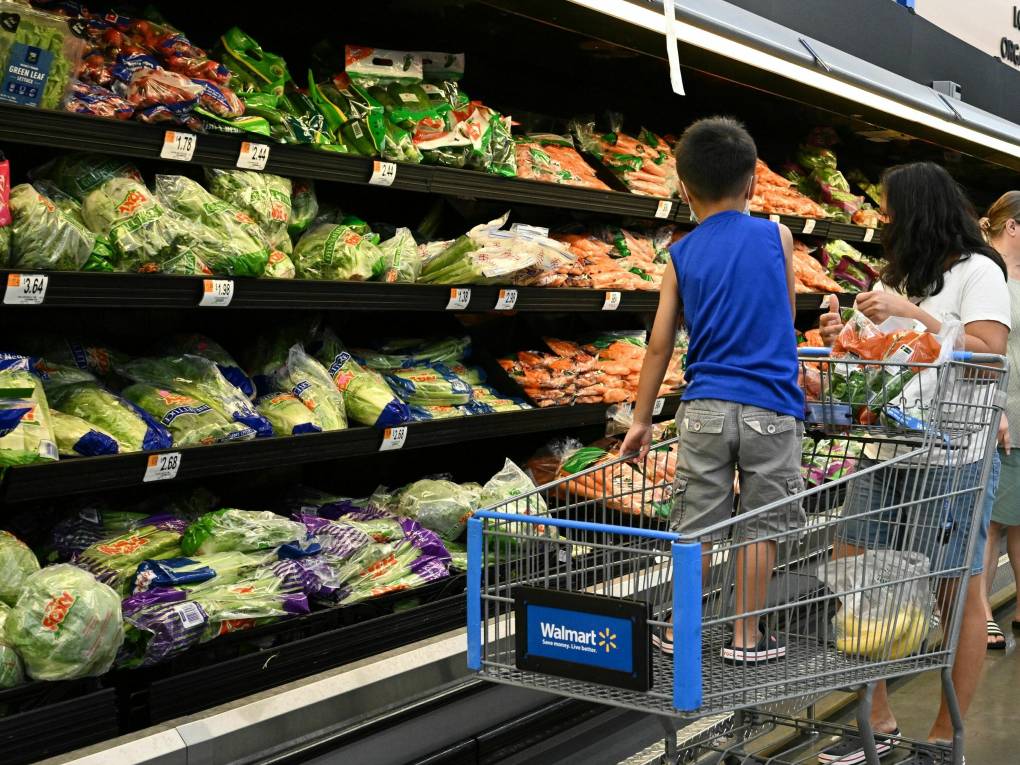 A small boy stands in a shopping cart next to a woman wearing a white shirt looking at vegetables in a grocery store.