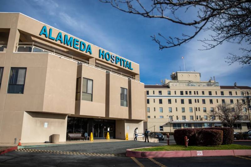 A beige building with "Alameda Hospital" written on it in blue lettering and a carpark in the foreground.