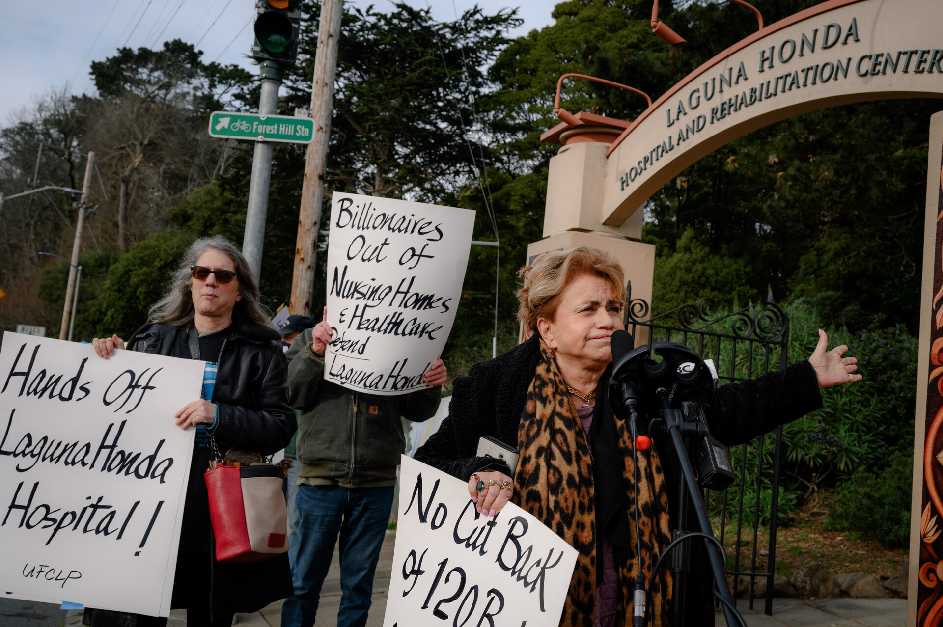 Three women with banners stand outside the Laguna Honda hospital entrance with protest signs in their hands.