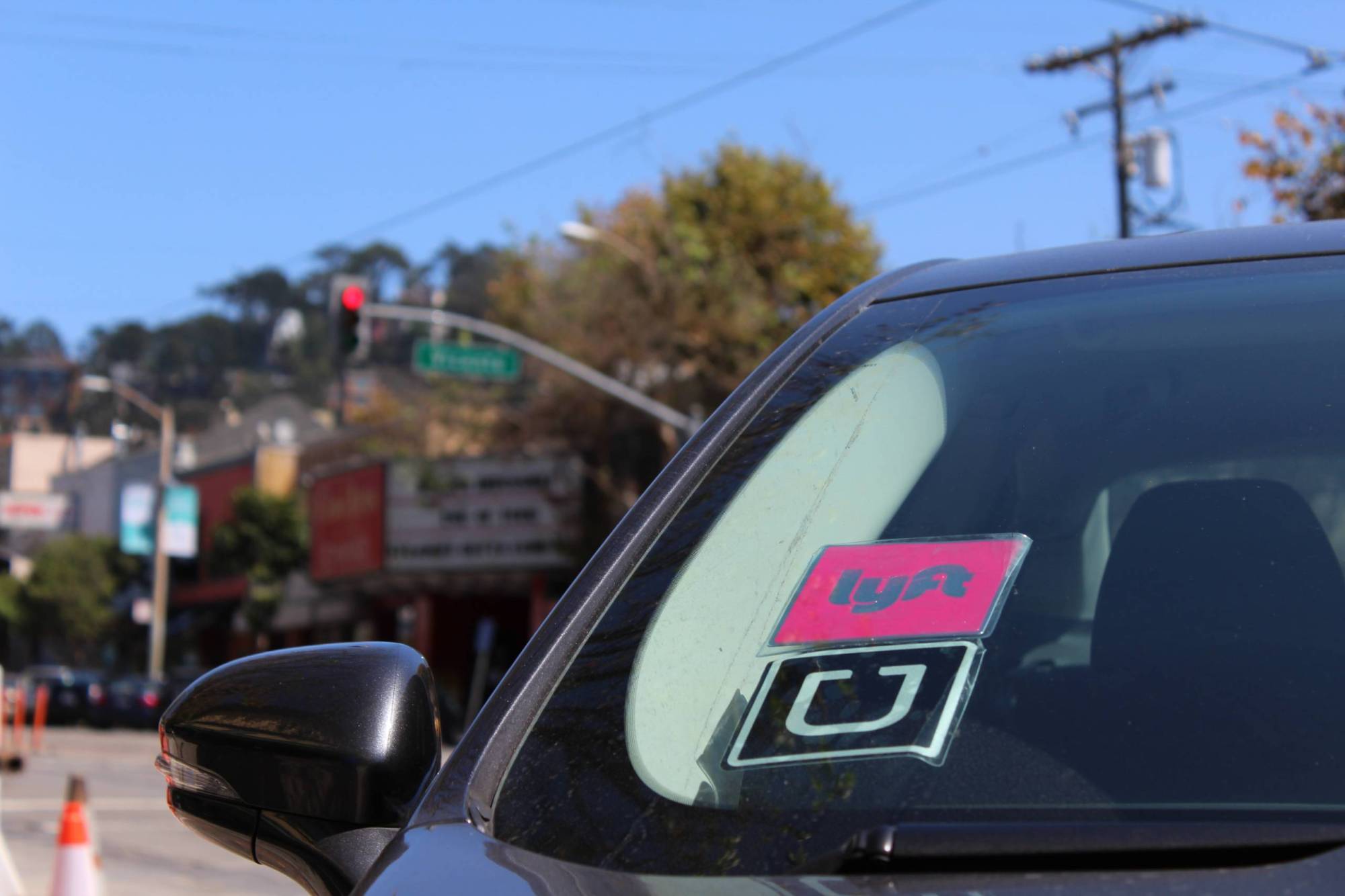 A closeup shot of a black vehicle with a pink Lyft sticker and a black and white Uber sticker on the left side of its windshield. The vehicle sits idle, waiting to pick up a customer.