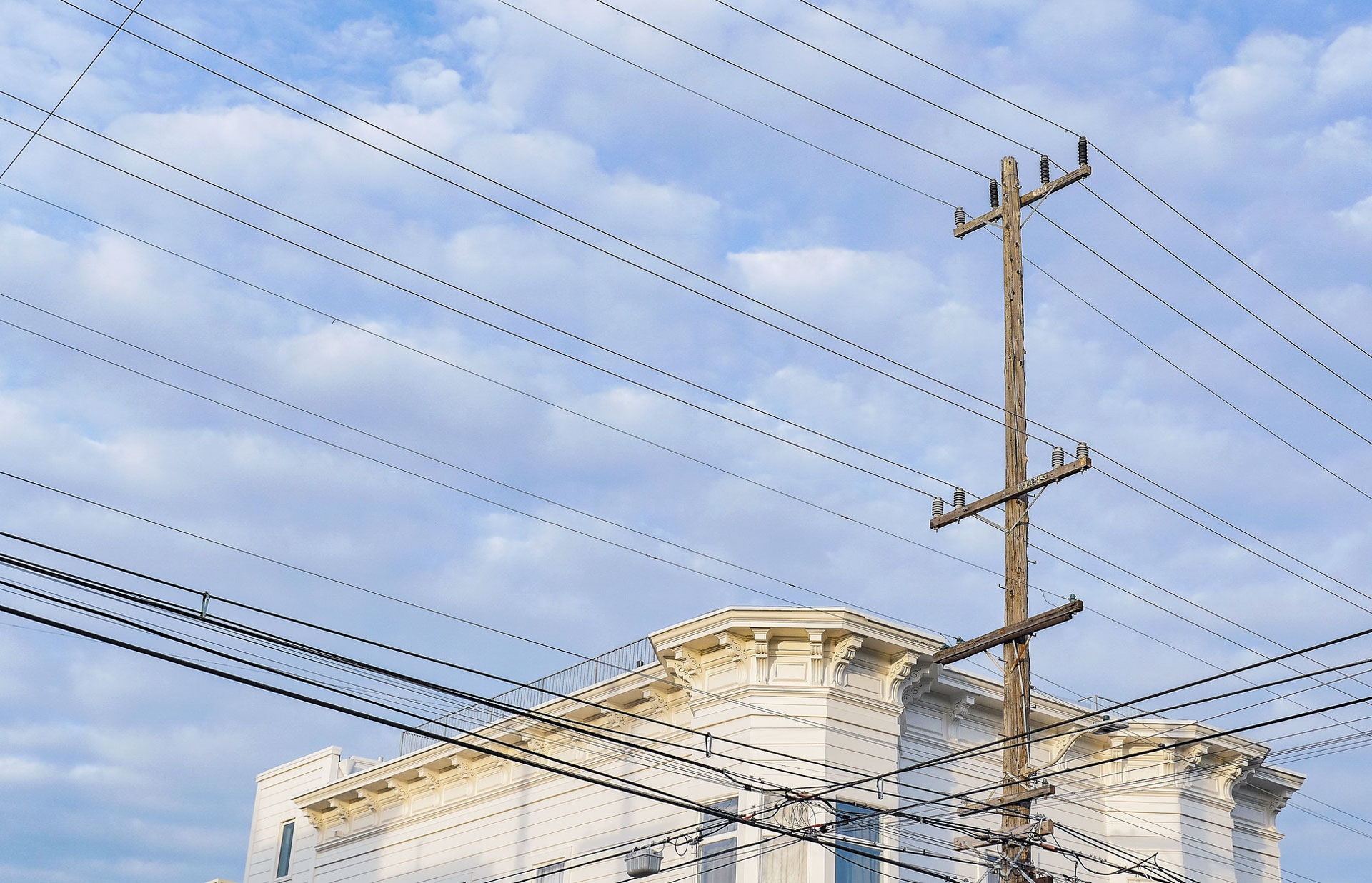 A photo shows primarily a wooden power pole with utility lines crossing the sky and the outline of the top of a building that looks like a San Francisco Victorian in the background.