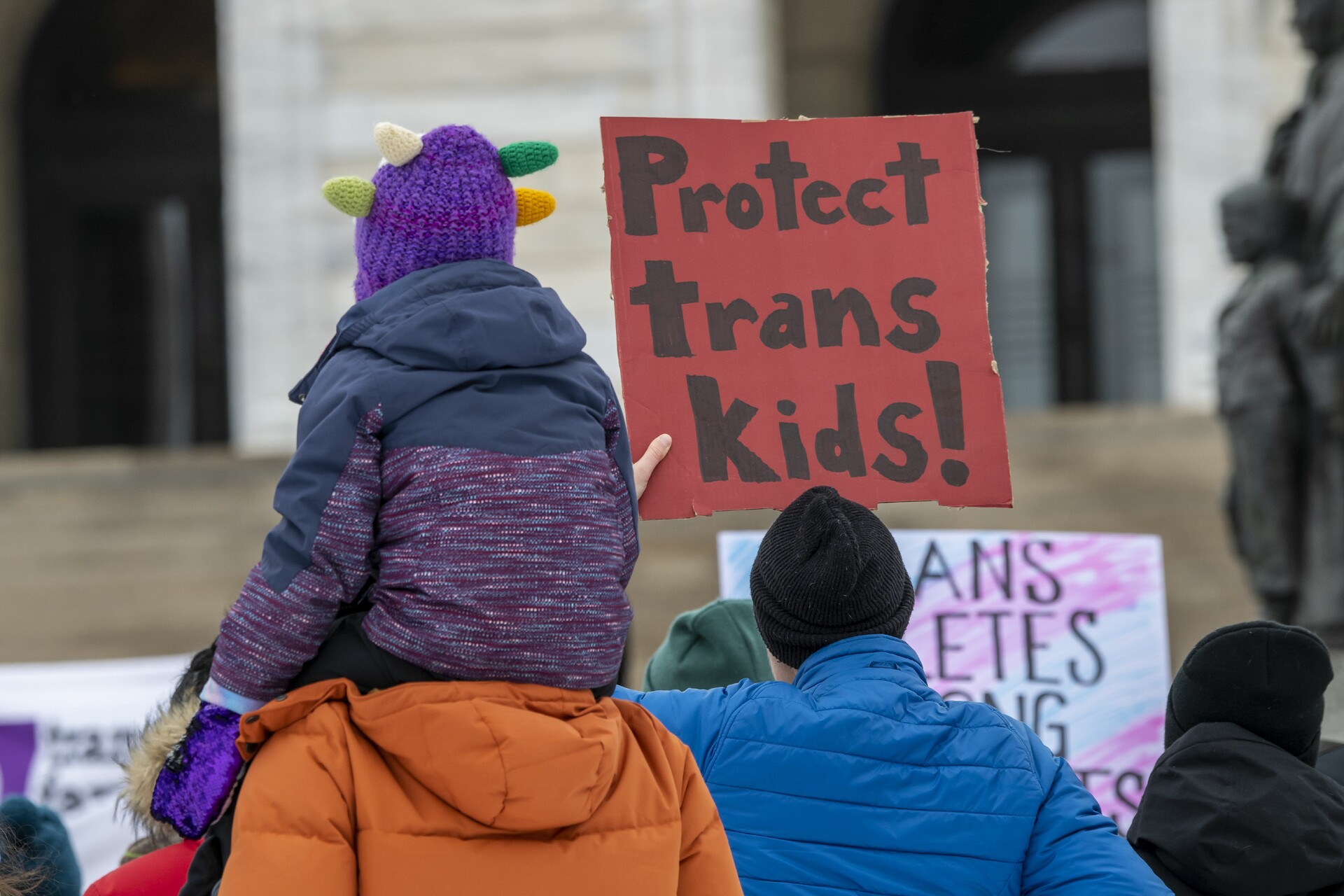 A family with two adults and a child on the shoulders of one adult looks on from behind, holding a sign that reads: "Protecting transgender children"