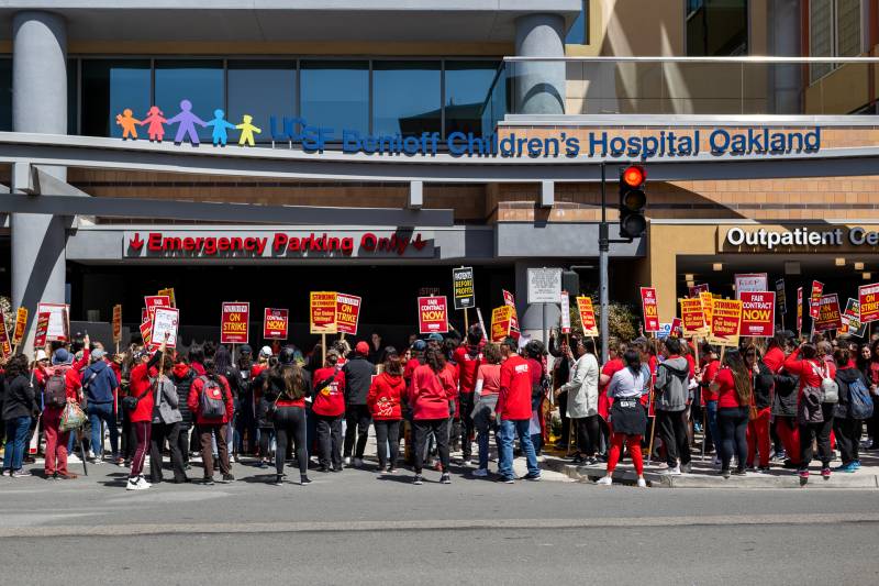 Hundreds of protesters in red T-shirts hold red and yellow picket signs outside of UCSF Benioff Children's Hospital Oakland.