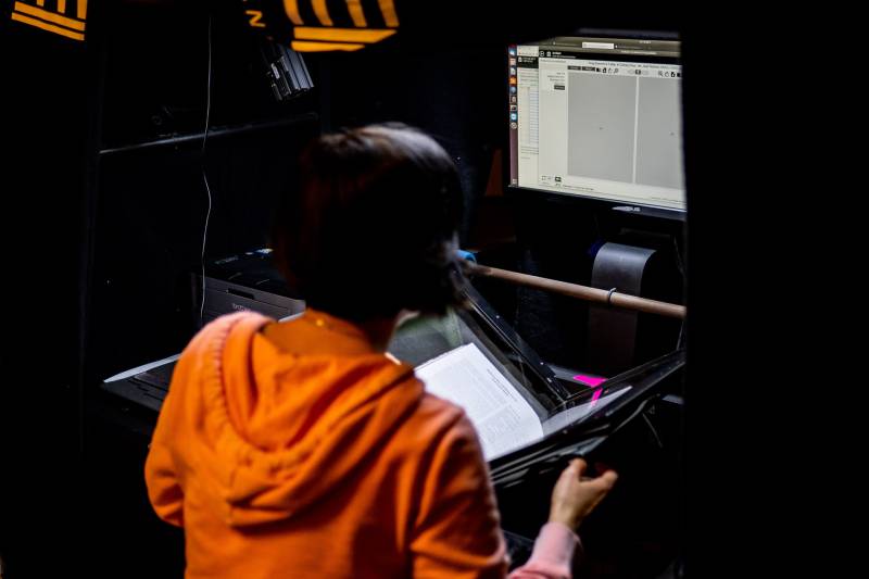 Someone wearing a bright orange hoodie sits at an archiving station, holding an open book and facing a computer screen.