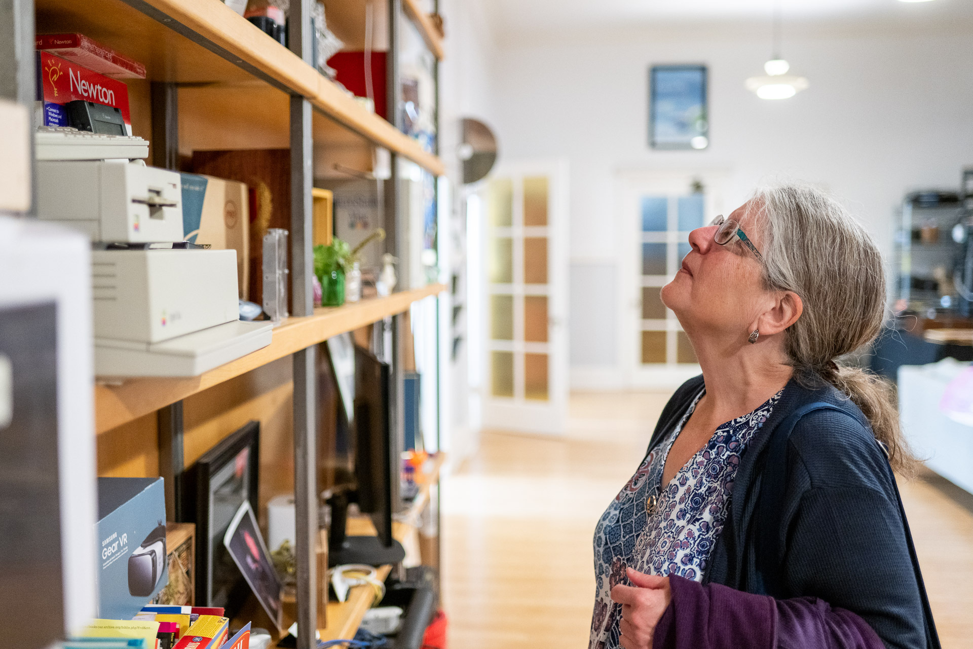 A middle-aged white woman with glasses and grey hair stands in front of what appears to be a large shelving unit full of memorabilia in a large, clean, well lit room