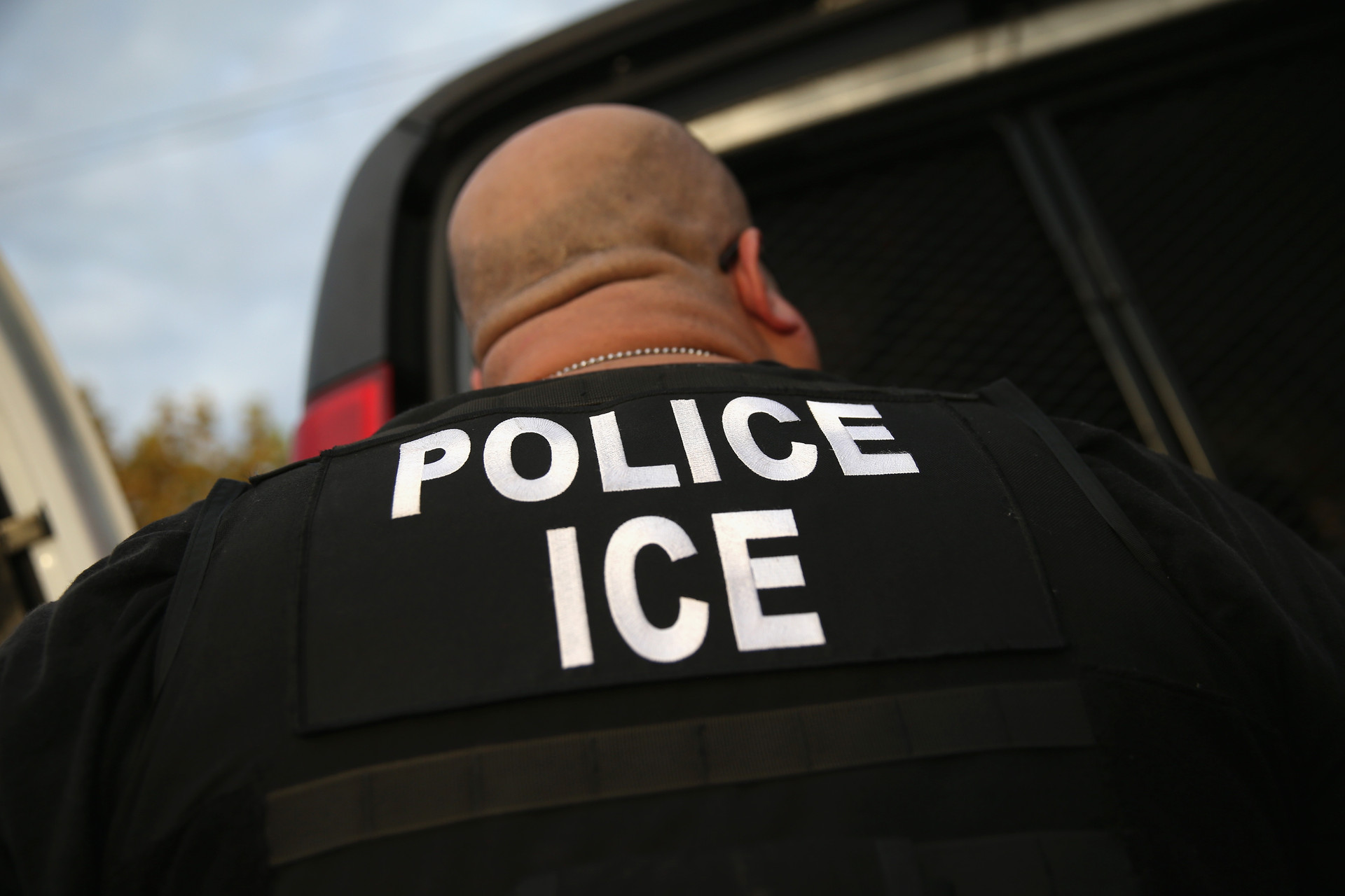 Detail shot of the back of a large man with a bald head and thick neck, wearing a black shirt with 'police ice' printed on it in all capital letters